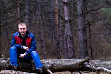 young attractive man dressed in a red jacket with blue sleeves sits on a log