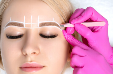 Woman Having Permanent Make-up Tattoo on her Eyebrows. Eyelash maker plucks eyebrows with tweezers....