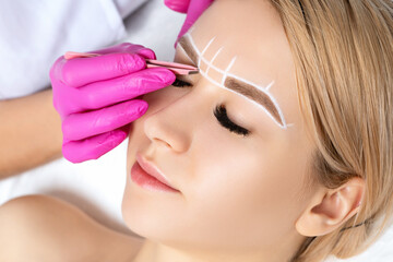 Woman Having Permanent Make-up Tattoo on her Eyebrows. Eyelash maker plucks eyebrows with tweezers....