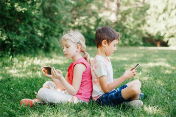 Boy and girl play games on smartphones outdoors. Kids digital gadget screen addiction. Children friends playing online sitting on grass in park. Contemporary problem of loneliness together.