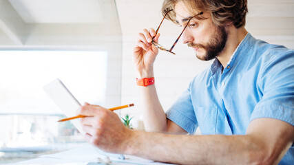 Handsome man with stubble and eyeglass working at home near big bright window in his own house....