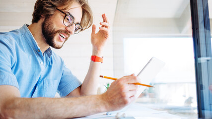 Handsome man with stubble and eyeglass working at home near big bright window in his own house. Remote workplace. Excited using tablet computer