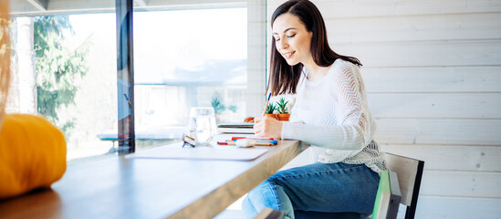 Handsome woman with long hair working at home near big bright window at her own house. Remote workplace. Smiling and drawing on the desk. Wide screen panoramic