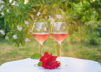 Romantic holiday composition. Two glasses of pink wine and red roses on white vintage table in the blooming garden