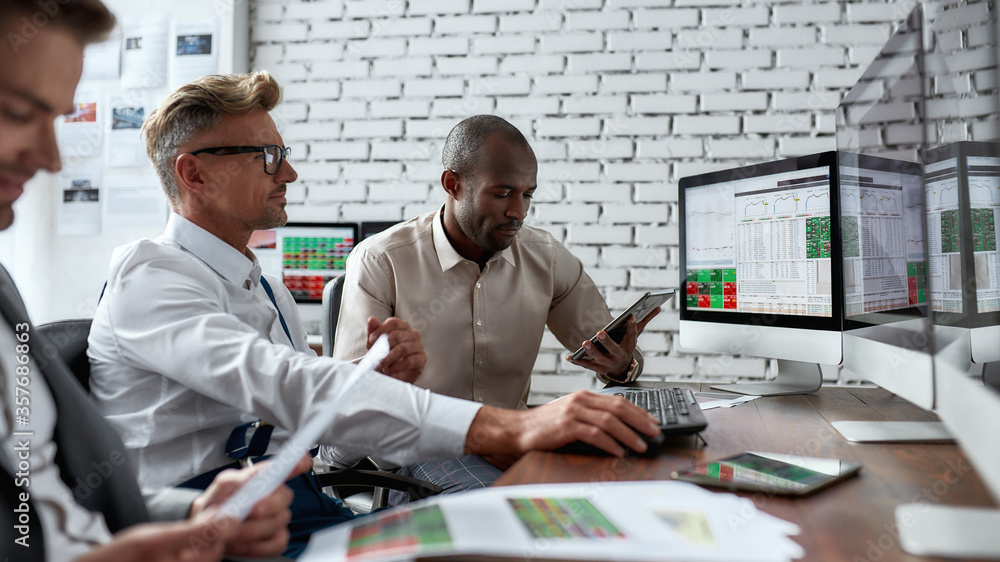 Wall mural Learn Together. Successful businessmen trading stocks. Stock traders looking at graphs, indexes and numbers on multiple computer screens. Three colleagues in modern office