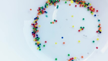 Falling sugar drops on a white plate. Multi-colored beads. Slow-motion. Abstract background with glass particles. - Powered by Adobe