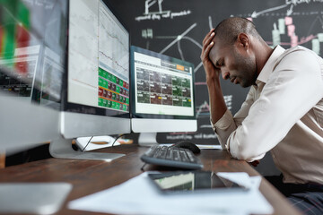 Have the wise choice. Portrait of stylish african businessman, trader sitting by desk in front of multiple monitors. He is getting disappointed while looking at current graphs