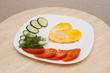 fried eggs in the form of a heart lie on a white plate, next to chopped dill, tomatoes and cucumbers