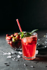 A refreshing summer drink made from strawberries and mint, juice, soda with ice cubes and slices of berries, in a beautiful glass with water droplets on a wooden board, dark background, vertical photo