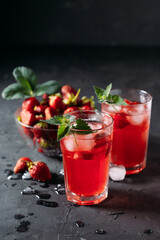 A refreshing summer drink made from strawberries and mint, juice, soda with ice cubes and slices of berries, in a beautiful glass with water droplets on a wooden board, dark background, vertical photo