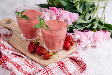 Strawberry smoothie with mint leaves in glasses on a wooden board with a beautiful serving of red napkin, berries and peony flowers on a light background.