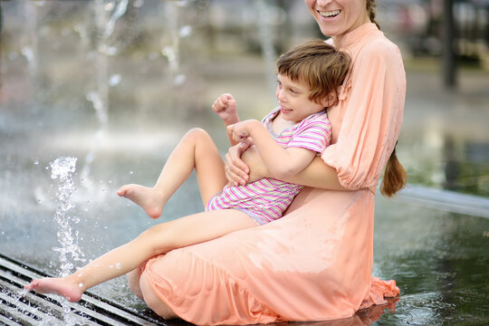 Portrait of beautiful disabled girl in the arms of his mother having fun on fountain of public park at sunny summer day. Child cerebral palsy. Disability. Inclusion.