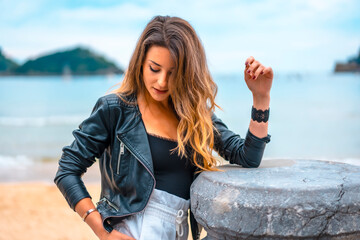 Lifestyle in the city with a blonde girl in white pants and a leather jacket. Pensive near the sea