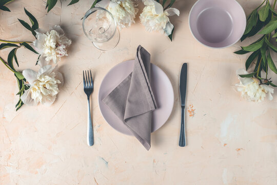 Flat lay composition with white peony flowers, empty plate with napkin and cutlery  on a light pink concrete background