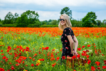 Fototapeta na wymiar Blonde girl in beautiful dress with suitcase in poppies field in summer time