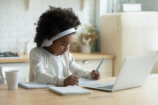 In kitchen schoolgirl do homework, focused little African girl wear headphones watch video lesson using laptop app, interested in on-line web virtual class studying from at home, homeschooling concept