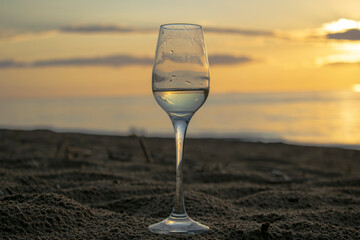 One glass of white wine. It stands on the sand against the backdrop of the sunset and the beautiful sky.