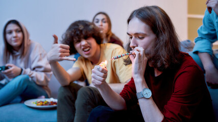 Great time. Young teenager, man lighting cannabis cigarette while sitting with excited friends on the couch at home. Young people smoking marijuana together, having fun