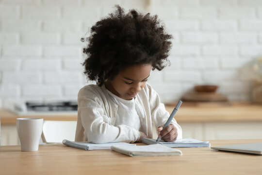 Clever focused little african ethnicity girl sitting at table in kitchen doing homework learn school subject hold felt-pen writing on workbook. Studying at home, homeschooling, self-education concept
