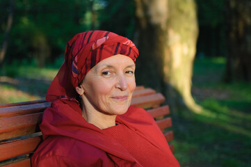 Female portrait of mature woman dressed in red clothes and sitting on the bench and enjoying a good weather and sunset in the park during a spring or autumn.