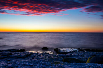 Obraz premium Vibrant colored summer sunset reflecting in ocean with endless horizon over deep blue ocean, silhouette of boulders that is laying in the foreground in shallow water at island of Gotland, Sweden