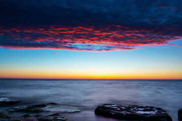 Obraz premium Vibrant colored summer sunset reflecting in ocean with endless horizon over deep blue ocean, silhouette of boulders that is laying in the foreground in shallow water at island of Gotland, Sweden