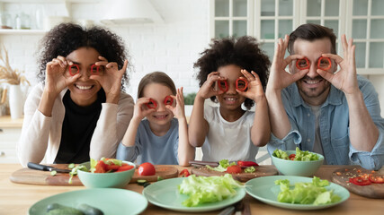 In kitchen couple and multi racial daughters prepare vegetable salad having fun make funny faces cover eyes with red paprika circles looking like eyewear, binoculars shape, cookery, family joy concept