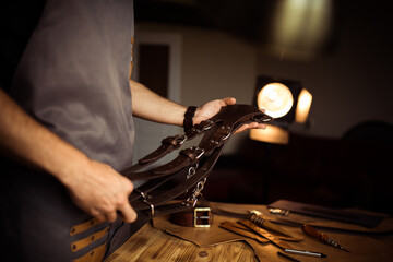 Working process of the leather belt in the leather workshop. Man holding photographer's belt for camera. Tool on wooden background. Tanner in old tannery. Close up master's arm