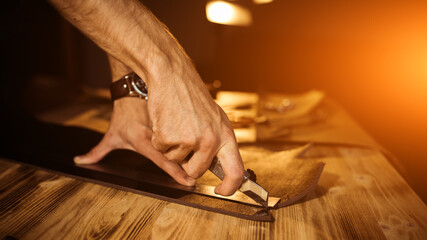 Working process of the leather belt in the leather workshop. Man holding tool. Tanner in old tannery. Wooden table background. Close up man arm. Warm Light for text and design. Web banner size