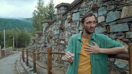 Adult man smoking cigarette with a fit of heart pain, standing along the road,  stone wall on...