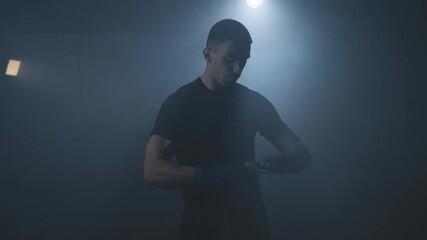 Male boxer wrapping hands with boxing tapes before fighting in smoky gym. Close-up of young athlete getting ready for fight. Sport concept in 4K, UHD. Man preparing.