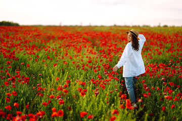 Young girl in hat posing in the poppy field. Portrait of curly girl walking in poppy field at sunset. Summertime.