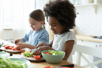 Obraz na płótnie Canvas African and Caucasian little girls best friends cooking together in modern kitchen. Multiracial cousins hold knives cutting vegetables on wooden board prepare healthy salad making surprise for parents