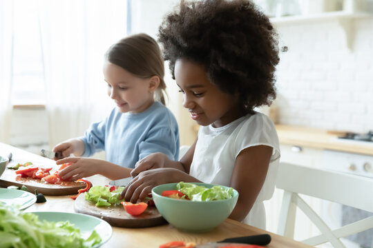 African and Caucasian little girls best friends cooking together in modern kitchen. Multiracial cousins hold knives cutting vegetables on wooden board prepare healthy salad making surprise for parents