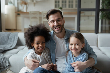 Father Day celebration, happy fatherhood multiracial family portrait concept. Caucasian stepdad sit on sofa hugs european mixed-race african daughters people smile pose for camera photo shoot at home