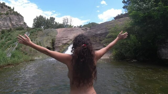 Nudist girl from the back raises her arms in a river and a waterfall in a natural landscape