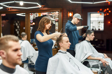 Getting haircut. Side view of professional barber girl holding scissors and comb in hands and cutting hair of young guy in the modern barbershop. Selective focus