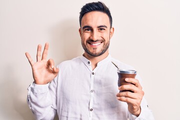 Young handsome man with beard drinking mate infusion beverage over white background doing ok sign with fingers, smiling friendly gesturing excellent symbol