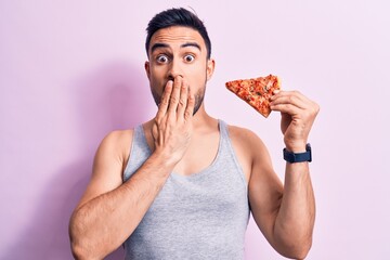 Young handsome man with beard eating slice of Italian pizza over isolated pink background covering...