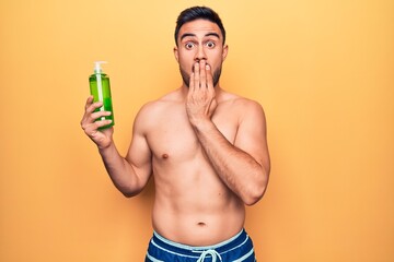 Fototapeta premium Handsome man with beard on vacation wearing swimwear holding bottle of aloe vera cream covering mouth with hand, shocked and afraid for mistake. Surprised expression