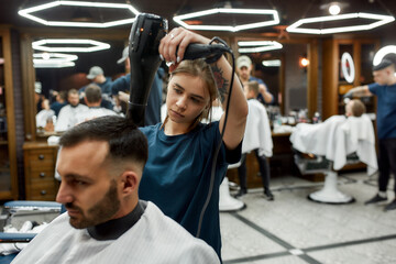 Modern barbershop. Young bearded handsome man sitting in barbershop chair while female barber...