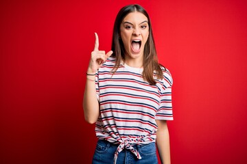 Young beautiful brunette woman wearing casual striped t-shirt standing over red background pointing finger up with successful idea. Exited and happy. Number one.
