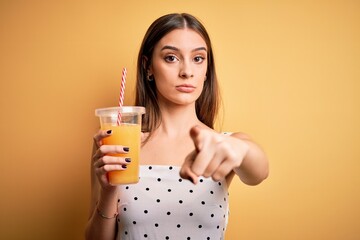 Young beautiful brunette woman drinking healthy orange juice over yellow background pointing with...