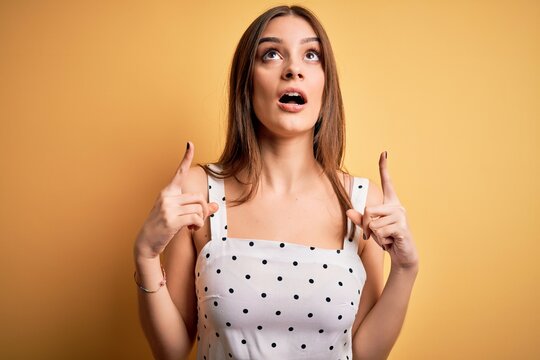Young beautiful brunette woman wearing casual dress standing over yellow background amazed and surprised looking up and pointing with fingers and raised arms.