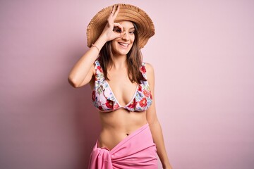 Young beautiful woman on vacation wearing bikini and summer hat over pink background doing ok gesture with hand smiling, eye looking through fingers with happy face.