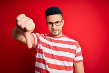 Fototapeta premium Young handsome man wearing casual striped t-shirt and glasses over isolated red background looking unhappy and angry showing rejection and negative with thumbs down gesture. Bad expression.