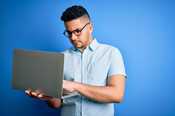 Young handsome businessman wearing glasses working using laptop over blue background with a confident expression on smart face thinking serious
