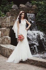 Fashion portrait of a beautiful bride. The bride in a white wedding dress stands against the backdrop of an artificial waterfall in a city park.