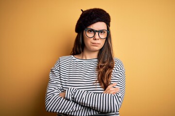 Young beautiful brunette woman wearing french beret and glasses over yellow background skeptic and nervous, disapproving expression on face with crossed arms. Negative person.