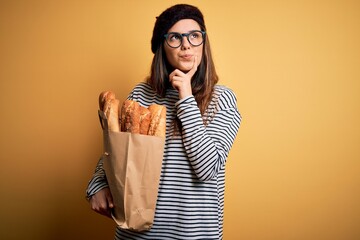 Young beautiful brunette french woman wearing beret holding paper bag with baguettes serious face thinking about question, very confused idea